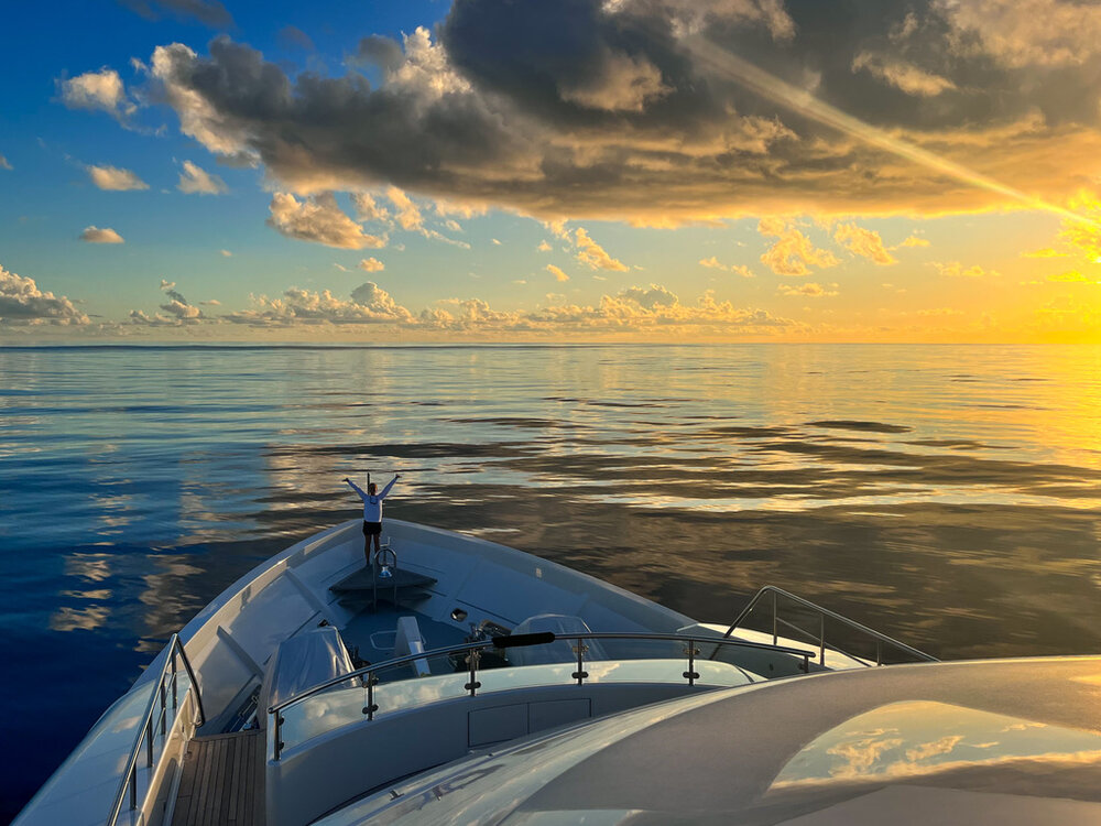 a zoomed out image of a girl standing on the bow of a yacht with a beautiful sunset over the very flat ocean